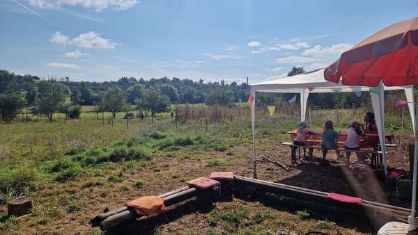 Ausblick über das Gelände mit Feuerstelle, Sonnenschirm und Kinder am Tisch.